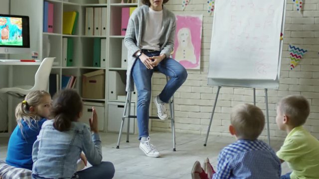 Tilt up of group of little boys and girls sitting on the floor in kindergarten classroom and listening to young female teacher