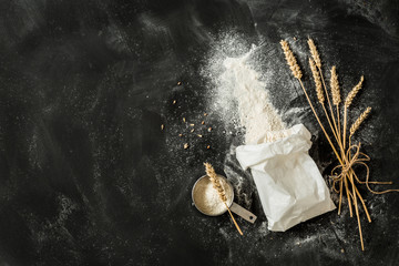Flour bag, ears of wheat and measuring cup on black background