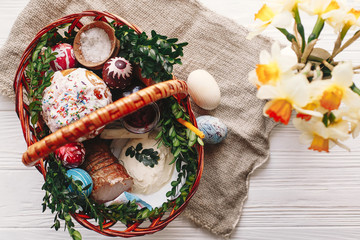 happy Easter concept. stylish basket with painted eggs, bread, ham,beets, butter on rustic wooden background with spring flowers and candle. easter food for blessing in church.