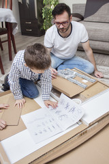Ojciec z synem klęczą na dywanie przed pudełkiem z elementami do samodzielnego złożenia i czytają instrukcje montażu