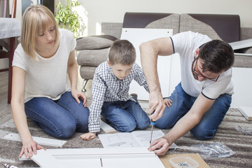 Rodzina skręca meble w salonie na dywanie. Ojciec wkręca śrubę wkrętakiem a mama i dziecko przyglądają się pracy. 
