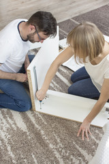 Kobieta i mężczyzna w jeansach i białych podkoszulkach siedzą na dywanie w salonie mieszkania i skręcają białe meble.  - obrazy, fototapety, plakaty