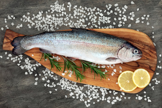 Rainbow trout healthy heart food on an olive wood board with rosemary herb, course sea salt and lemon on marble background. Very high in omega 3 fatty acid. 