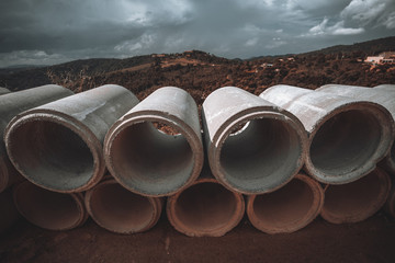 Wide-angle view of the two rows of the cement or concrete soil-pipes laying on the ground, used for the creation of the sewerage systems and the aqueducts, the stormy overcast sky in the background