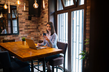 Freelance young woman sitting in the cafeteria with laptop and using mobile phone