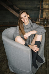 Portrait of a beautiful woman sitting cross-legged on the grey armchair wearing gaiters and long grey knitted cozy sweater. Happy girl in dayligth studio home posing. Closeup.