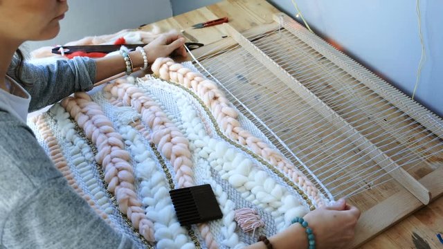 Weaving on a loom. Closeup woman's hands running on a loom. Threading the shuttle through the strands of frame and fasten the yarn with scallop.