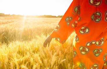 African woman in traditional clothes walking with her hand on a field of crops at sunset or sunrise