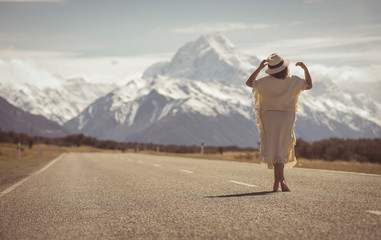 A boho hippy lady in white dress with hat stands on road with aoraki snow mountain background in New Zealand. idea for travel, destination.