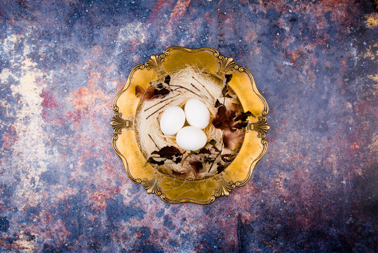 White eggs in stylish straw nest decorated with leaves and feathers on the old golden platter. Old rusty background. Flat lay, top view. Easter concept.