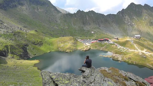 Young man sitting on the rock takes photos of the lake surrounded by mountains