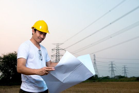 An engineer wear yellow hard hat and suite hold blueprint in hand standing on field looking at blueprint