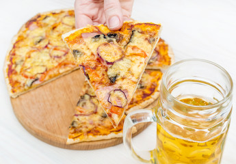 Hand holding piece of pizza over table with pizza and glass of beer.