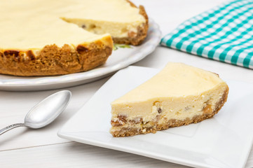 Piece of cheesecake on saucer with whole cheesecake and kitchen towel on the table.