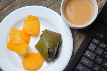 A cup of coffee, crispy potato chips and koci the malay glutinous rice ball served in banana leaf on white plate over wooden table