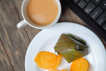 A cup of coffee, crispy potato chips and koci the malay glutinous rice ball served in banana leaf on white plate over wooden table