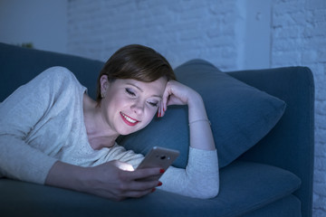 pretty and happy red hair woman on her 20s or 30s lying on home couch or bed using mobile phone late at night smiling in internet addiction