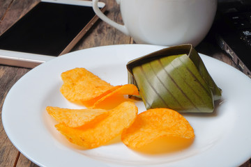 A cup of coffee, crispy potato chips and koci the malay glutinous rice ball served in banana leaf on white plate over wooden table