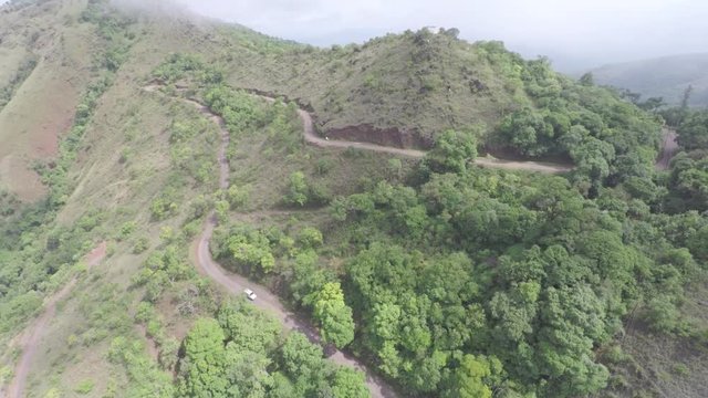 An aerial view of a car passing through canopies of trees on a mountain. 