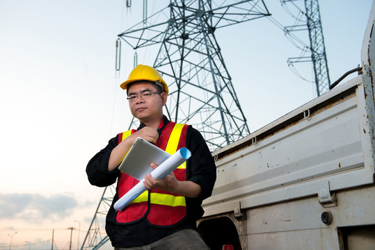 Electrical engineer working. Electrician holding blueprint and tablet at high voltage power pylon against blue sky