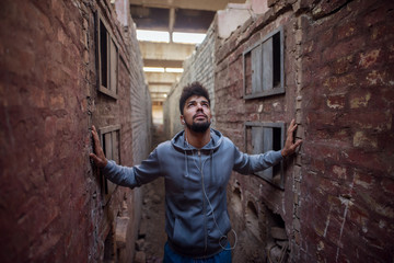 Fototapeta na wymiar Portrait of focused motivated afro-american young handsome sportive man with earphones standing inside of the abandoned place in the middle of two walls and thinking while looking up.