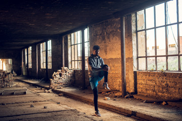 Portrait of active afro-american young attractive athletic man with earphones standing and doing stretching workout with raised leg inside of the abandoned place.