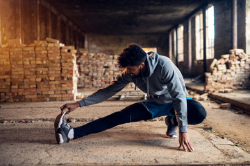Close up portrait of active afro-american young attractive athletic man doing full leg stretching workout inside of the abandoned place.
