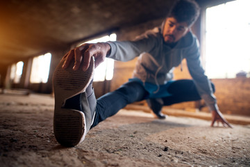 Close up portrait of active afro-american young attractive athletic man doing full leg stretching workout inside of the abandoned place.