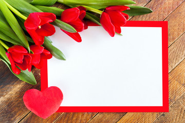 White blank sheet in red frame, red tulips and plush heart on rustic wooden background.