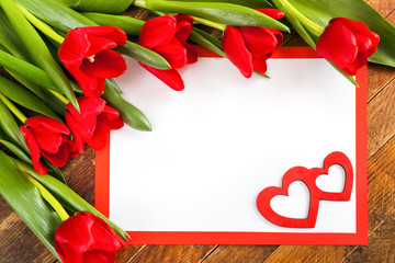 White blank sheet, red frame, red tulips and two red hearts on  diagonal wooden background.