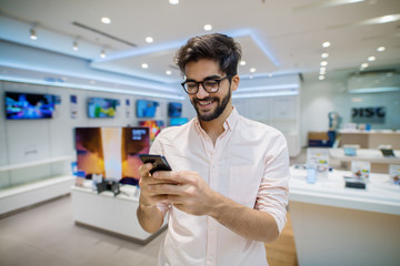 Close up portrait view of satisfied excited happy smiling young student bearded man with eyeglasses looking at mobile in a tech store.