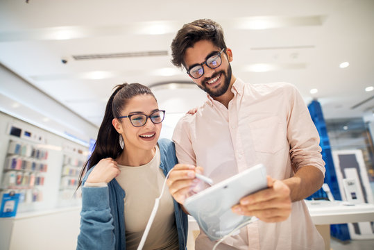 Low view of young cute stylish smiling couple testing the new model of a tablet with a pencil in the tech store.