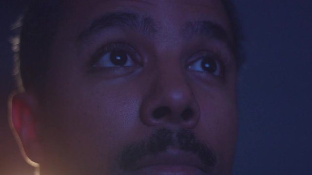 surprised mixed race man watching tv in darkness with different color light from tv on face. Eyes closeup