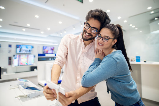 Portrait view of young cute love couple standing hugged in front of the desk with tablets and testing selfie operation in the tech store.