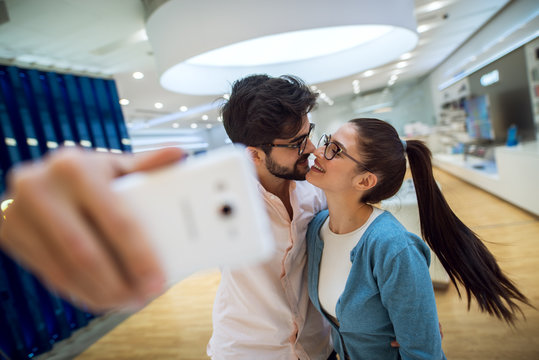 Close up focus view of a romantic adorable attractive young student love couple taking a selfie before kiss with new mobile in a tech store.