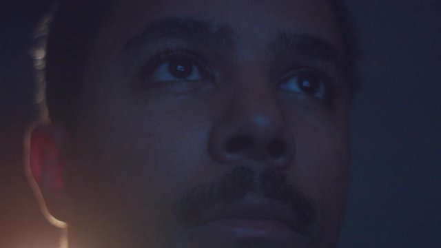 upset boring mixed race man watching tv in darkness with different color light from tv on face. Eyes closeup