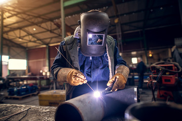 Close up portrait view of professional mask protected welder man in uniform working on the metal sculpture at the table in the industrial fabric workshop in front of few other workers. - obrazy, fototapety, plakaty