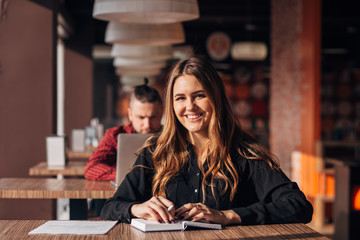 young woman smiling at camera while working remotely in coffee shop. Positive female with notepad