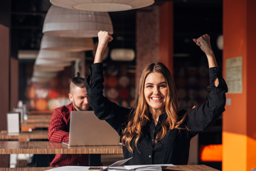 cheerful young businesswoman keeping arms raise celebrating success in cafe