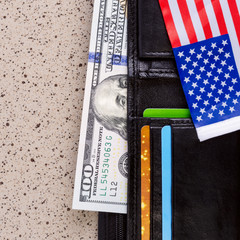 One hundred dollars banknote reach out out of a black old purse. And also part of the American flag. Square.