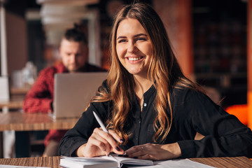 young woman smiling at camera while working remotely in coffee shop. Positive female with notepad