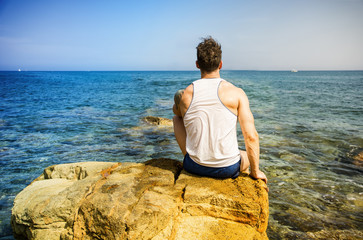 Handsome muscular man on the beach sitting on rocks, looking at camera