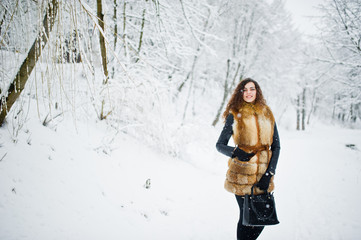 Elegance curly girl in fur coat and handbag at snowy forest park at winter.