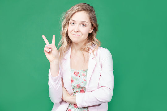 Fashionable female model with wavy hair, wears trendy colorful dress, round lips, gestures against green background, feels like star. Beautiful blonde woman shows peace sign. Body language concept.