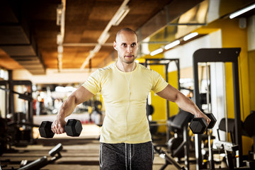Fototapeta na wymiar Portrait of strong muscular active healthy young man raising plastic dumbbells with open arms and listening music in the gym.