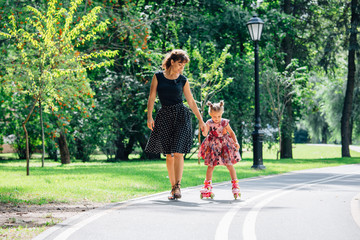 Mother teaching daughter to ride on rollers. Cheerful young woman and cute little daughter ride on...