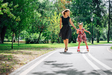 Mother teaching daughter to ride on rollers. Cheerful young woman and cute little daughter ride on...