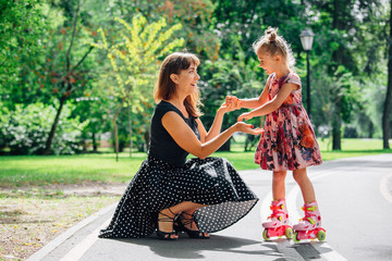 Mother teaching daughter to ride on rollers. Cheerful young woman and cute little daughter ride on...
