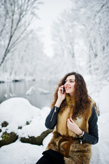 Fototapeta na wymiar Elegance curly girl in fur coat with mobile phone at snowy forest park at winter.
