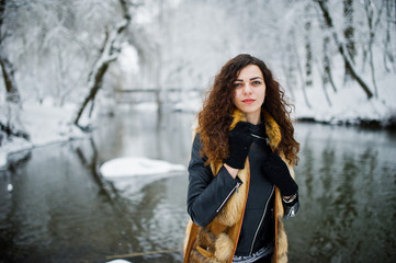 Elegance curly girl in fur coat at snowy forest park agasinst frozen river at winter.
